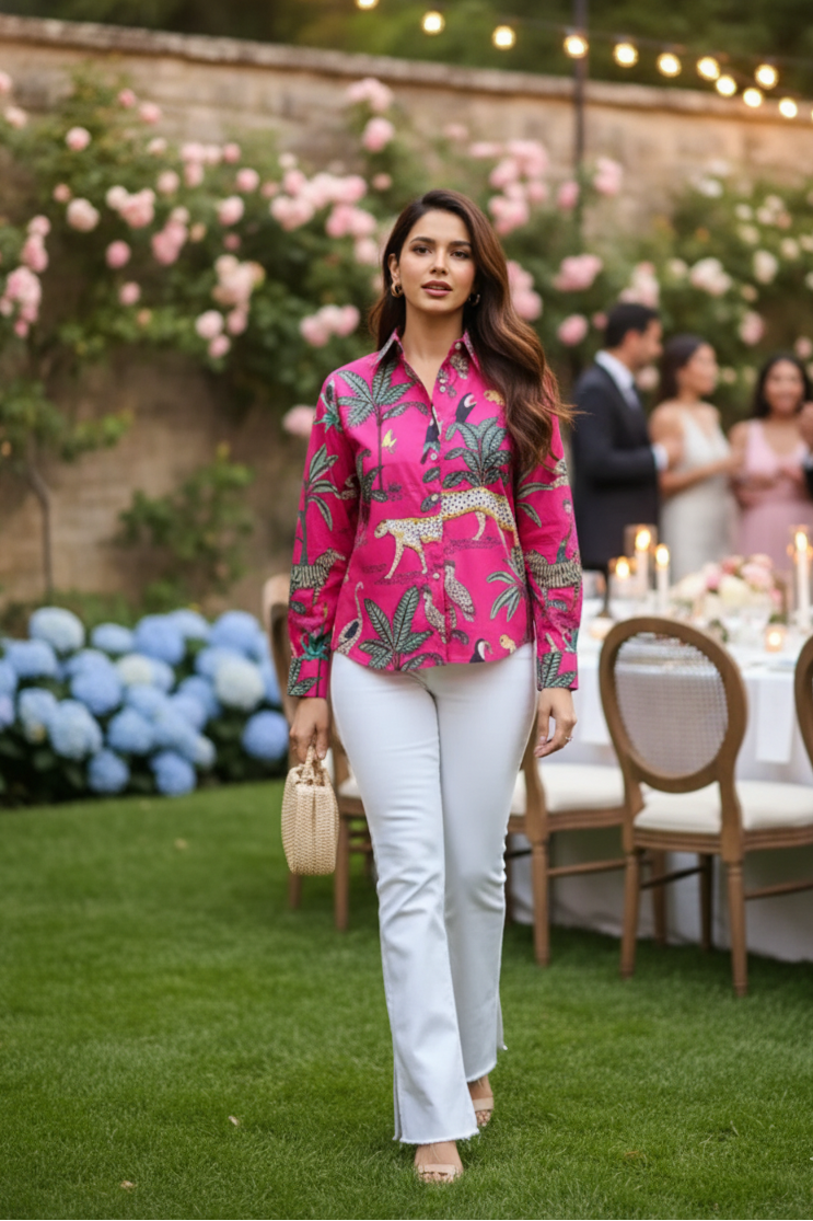 Woman in a pink floral blouse and white pants standing outdoors with floral decorations and people in the background.