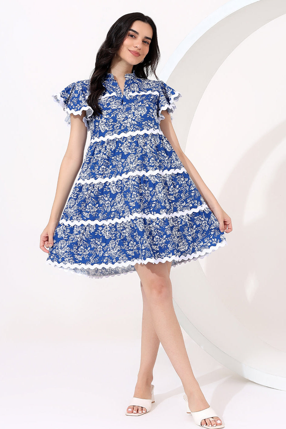 Woman in a blue floral dress standing in a garden with flowers and a bench in the background.