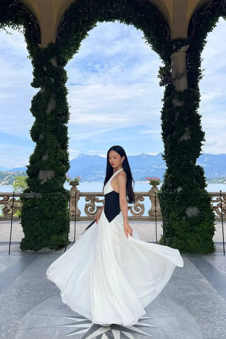 Woman in a black and white dress holding a black handbag in an outdoor setting with greenery and a building in the background.