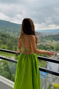 A woman stands on a balcony wearing a bright lime green backless maxi dress, looking out over a scenic mountain valley with green hills and buildings under a cloudy sky.