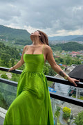 A woman wearing a bright lime green strapless maxi dress stands on a balcony with a scenic mountain and lake view in the background, looking upward under a cloudy sky.