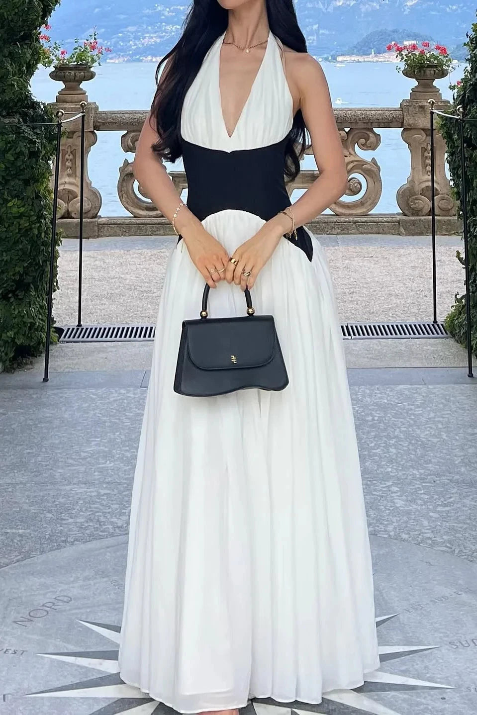 Woman in a black and white dress holding a black handbag in an outdoor setting with greenery and a building in the background.