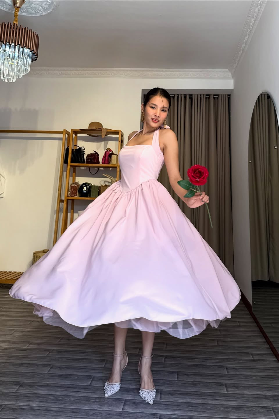 Woman in a pink dress holding a red rose in a room with a chandelier and shelves.
