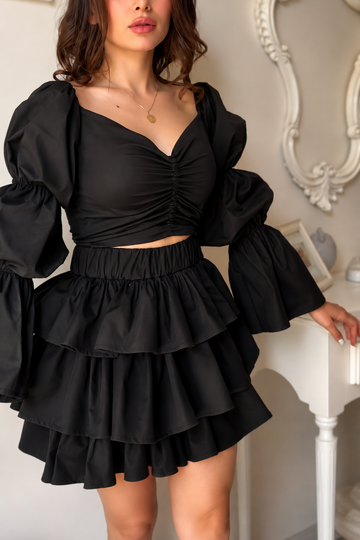 A woman modeling a black two-piece outfit consisting of a ruched sweetheart crop top with dramatic puffed sleeves and a matching tiered ruffled mini skirt, set against an elegant indoor background with a white ornate mirror.