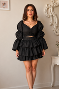 A woman modeling a black two-piece outfit consisting of a ruched sweetheart top with dramatic puffed sleeves and a matching tiered ruffle mini skirt, paired with black strappy heels in an elegant indoor setting.