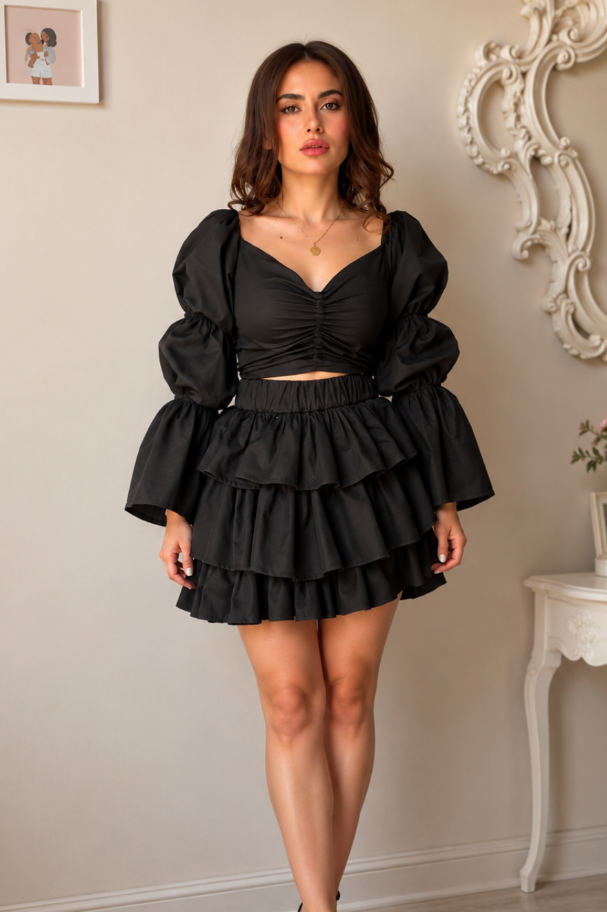 A woman modeling a black two-piece outfit consisting of a ruched sweetheart top with dramatic puffed sleeves and a matching tiered ruffle mini skirt, paired with black strappy heels in an elegant indoor setting.