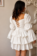 A woman shown from the back wearing a white tiered mini dress with puffed sleeves and a lace-up back, standing indoors against a beige wall with ornate wall decor.