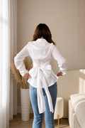 A woman viewed from behind wearing a white long-sleeve wrap blouse tied at the waist and blue jeans, standing in a softly lit bedroom with beige walls, curtains, and a bed.