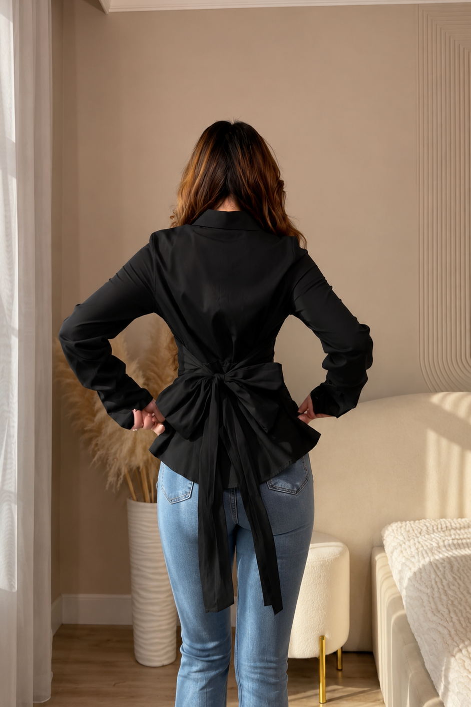 A woman viewed from behind wearing a black long-sleeve wrap-style blouse with a large tied back and light blue jeans, standing in a modern beige-toned bedroom or living space.