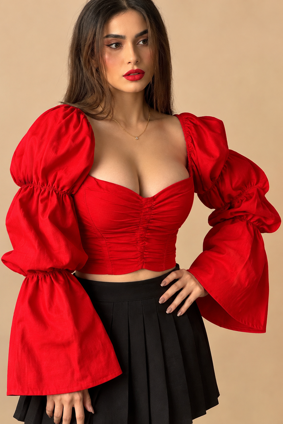 A woman modeling a bright red cropped bustier top with dramatic puffed bell sleeves and a black pleated skirt, set against a solid beige studio background.