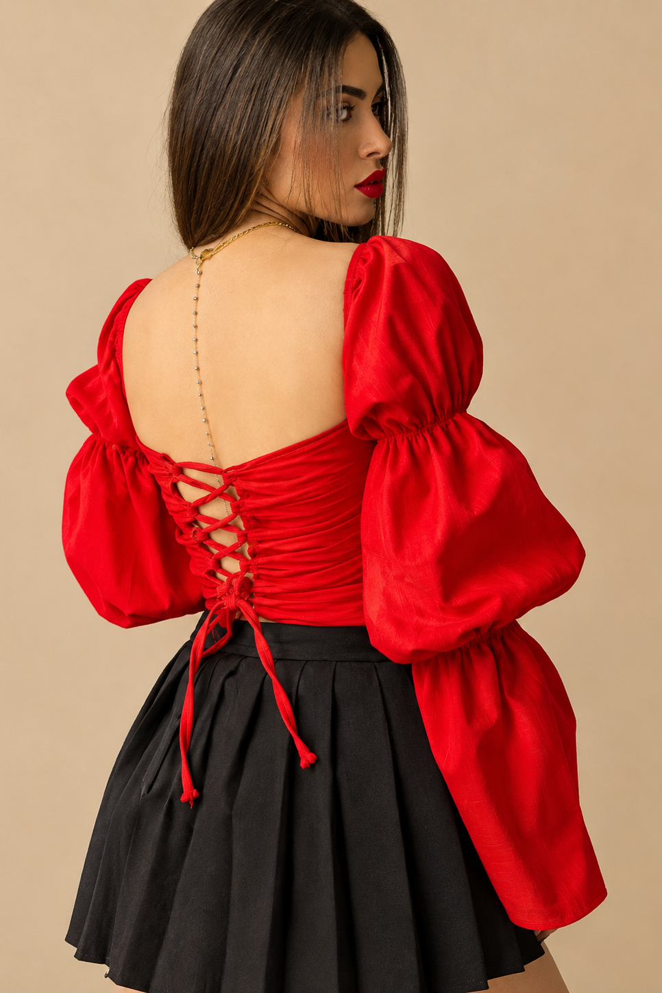A woman modeling a bright red cropped bustier top with dramatic puffed bell sleeves and a black pleated skirt, set against a solid beige studio background.