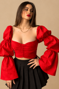 A woman modeling a vibrant red off-the-shoulder crop top with dramatic puffed bell sleeves and a black pleated skirt, set against a solid beige studio background.