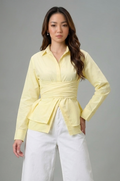 A model wearing a pale yellow long-sleeve peplum wrap blouse and white wide-leg cropped pants with fringe, paired with brown strappy heels, against a solid gray studio background.