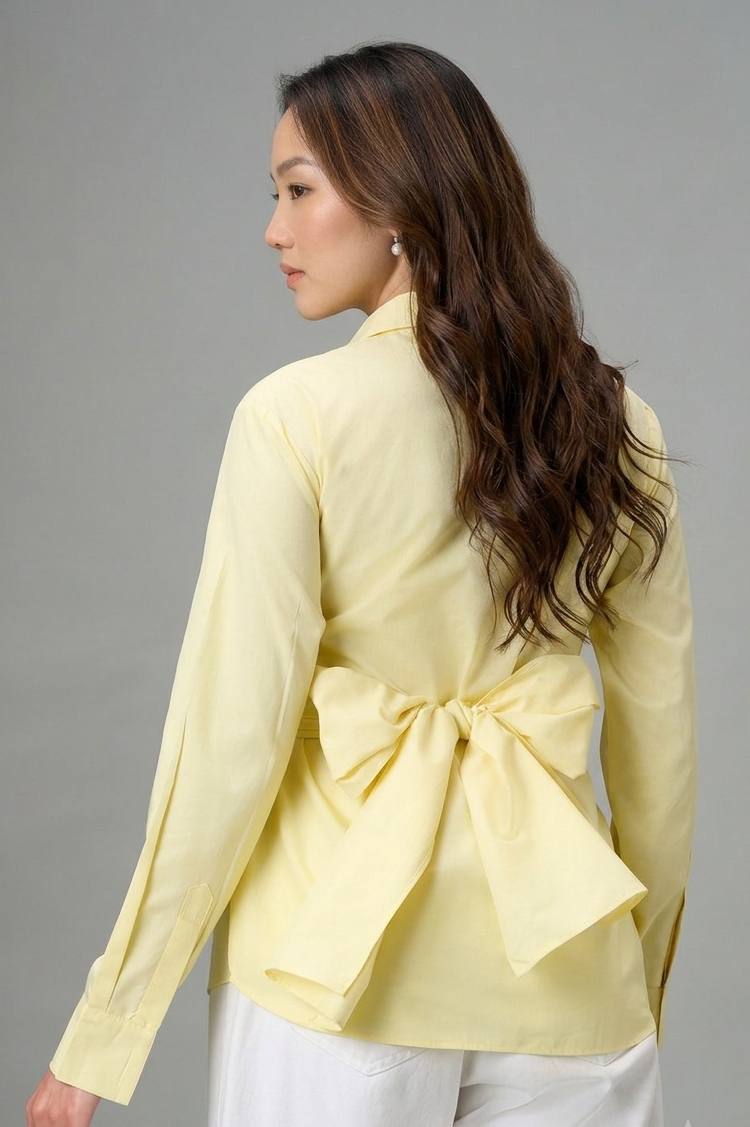 A model wearing a pale yellow long-sleeve peplum wrap blouse and white wide-leg cropped pants with fringe, paired with brown strappy heels, against a solid gray studio background.