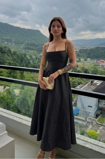A woman wearing a black spaghetti-strap midi dress stands on a balcony overlooking a scenic mountain landscape, holding a small beige clutch.