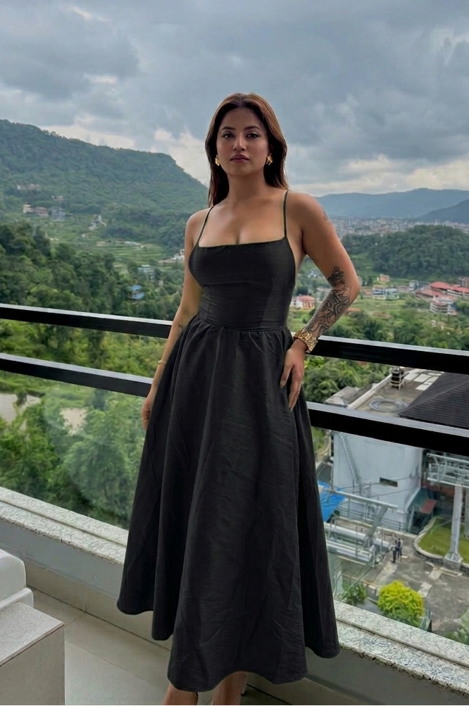 A woman wearing a black spaghetti-strap midi dress stands on a balcony with a scenic mountain and lake view in the background.