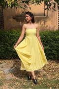 Woman in a yellow dress standing outdoors with greenery and a textured wall in the background