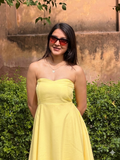 Woman in a yellow dress and sunglasses standing in front of a textured wall with greenery.