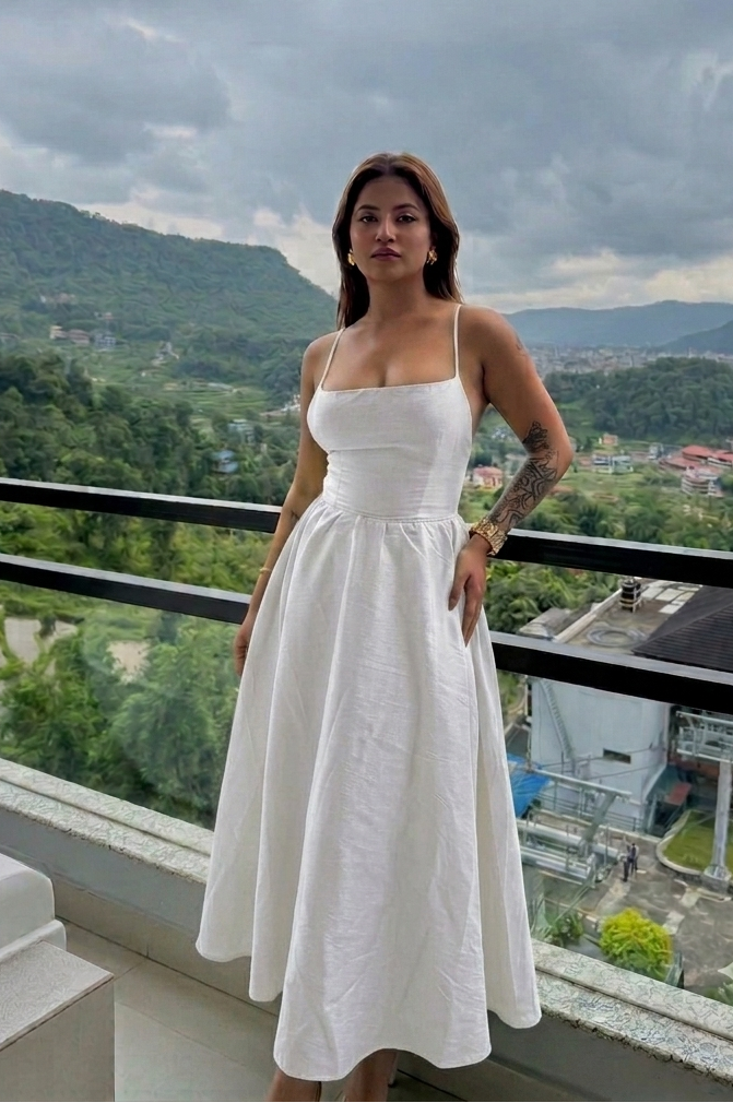 A woman wearing a white midi dress with spaghetti straps and a fitted waist stands on a balcony overlooking a lush green mountain valley under a cloudy sky.