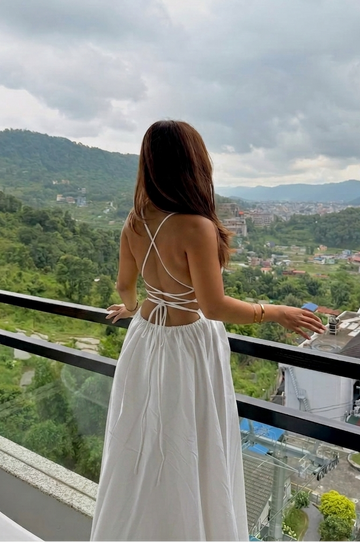 A woman stands on a balcony wearing a white maxi dress with a strappy back design, looking out over a lush green mountain town under a cloudy sky.