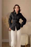 A woman stands in a bedroom setting wearing a black satin-like long-sleeve button-up shirt with a self-tie waist belt and white wide-leg trousers.