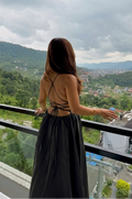 A woman stands on a balcony with a scenic mountain valley view, wearing a black backless maxi dress with strappy detailing on the back.