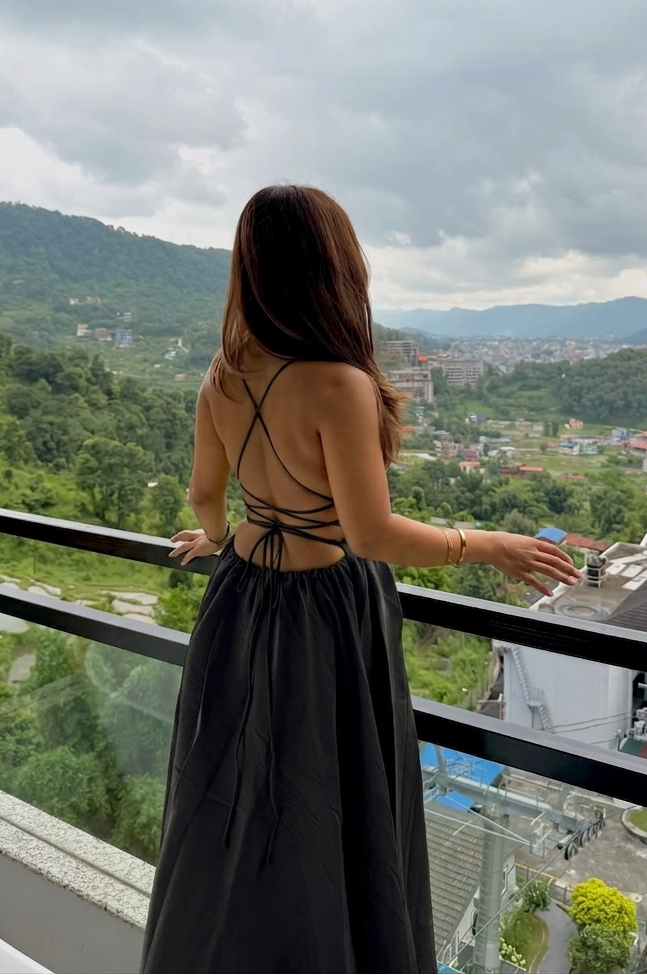 A woman stands on a balcony with a scenic mountain valley view, wearing a black backless maxi dress with strappy detailing on the back.