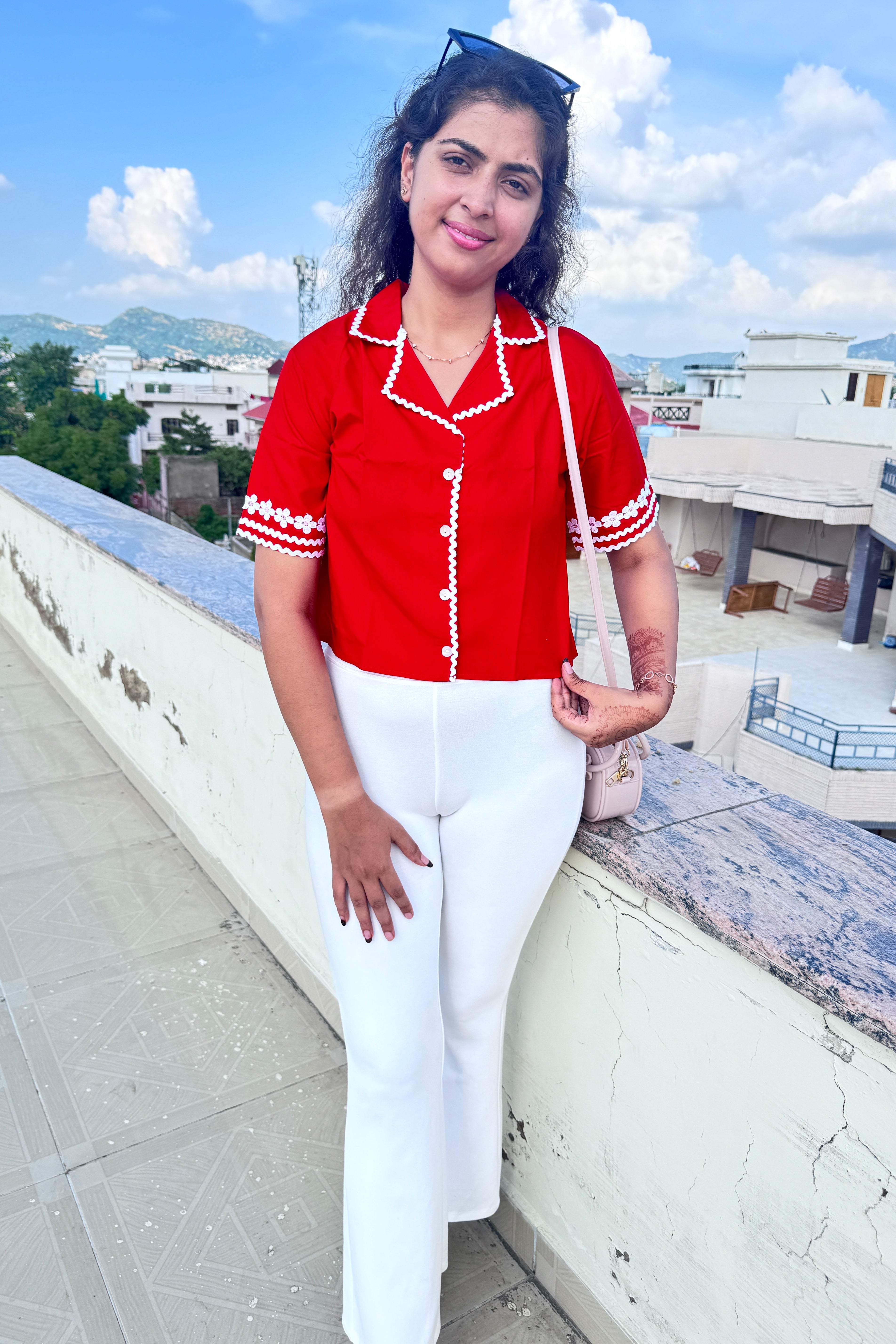 Woman in a red top and white pants posing outdoors with buildings and sky in the background