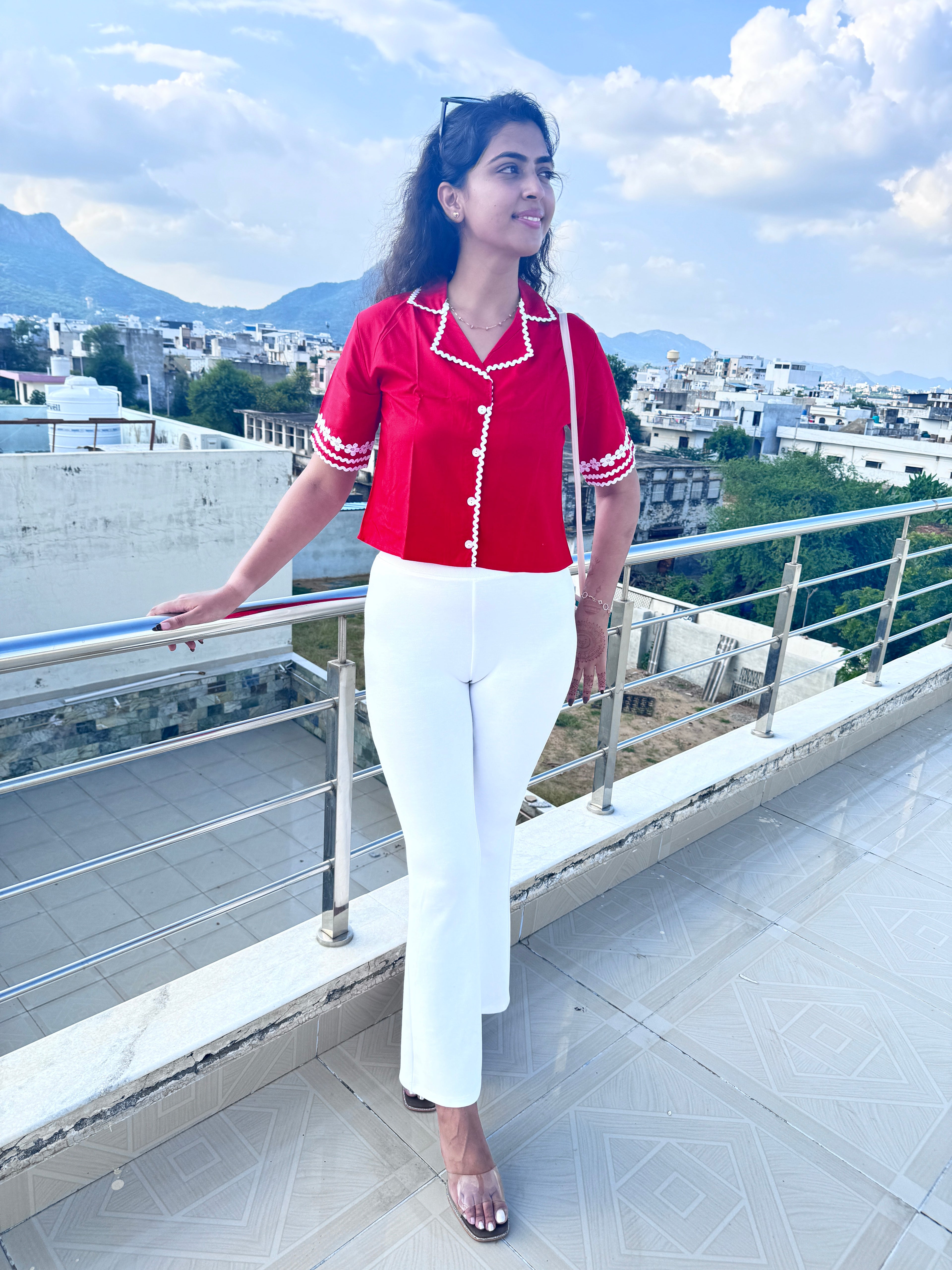 Person wearing a red top and white pants on a balcony with a cityscape view.