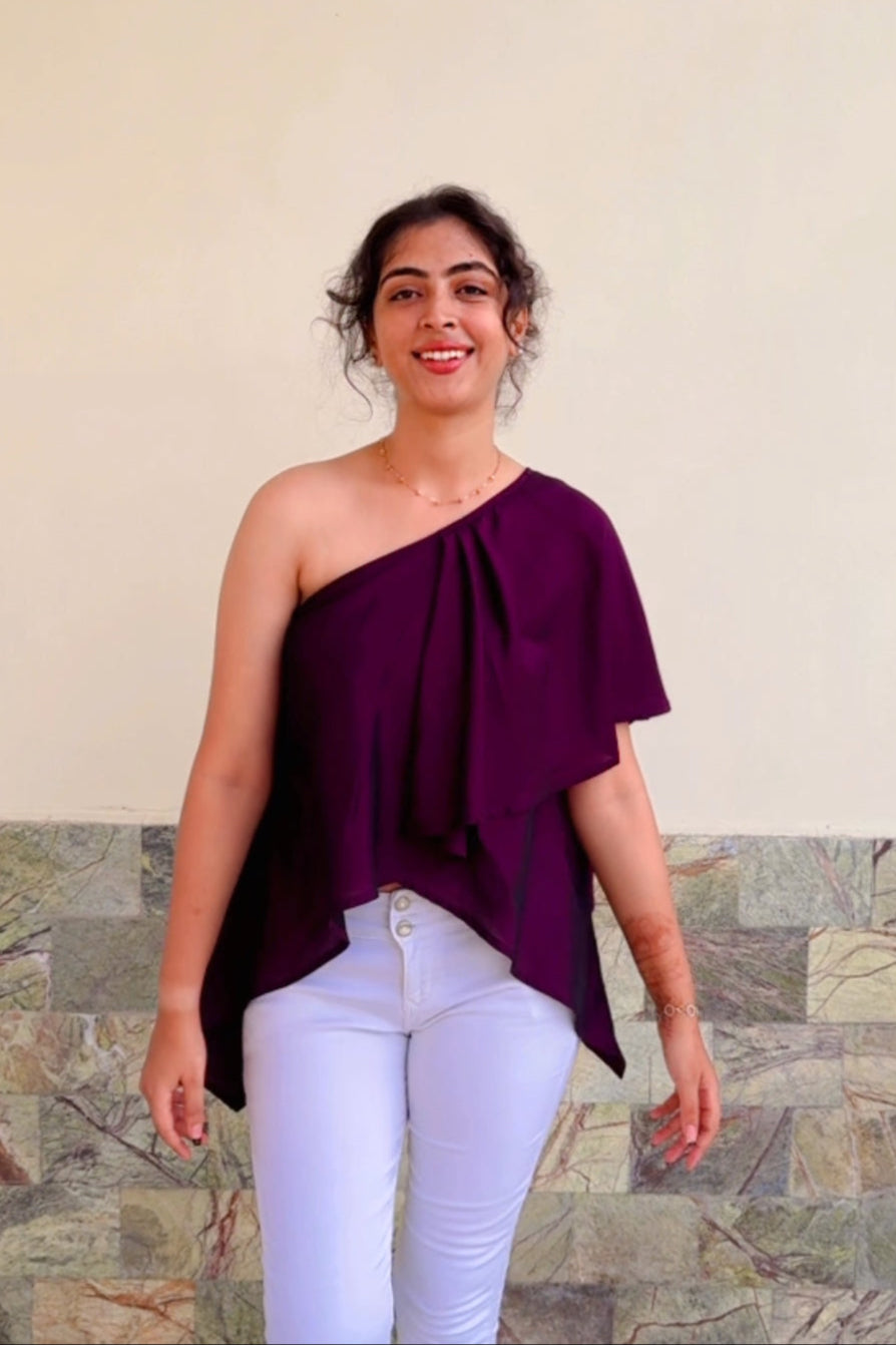 Woman wearing a one-shoulder purple top and white pants against a beige wall.