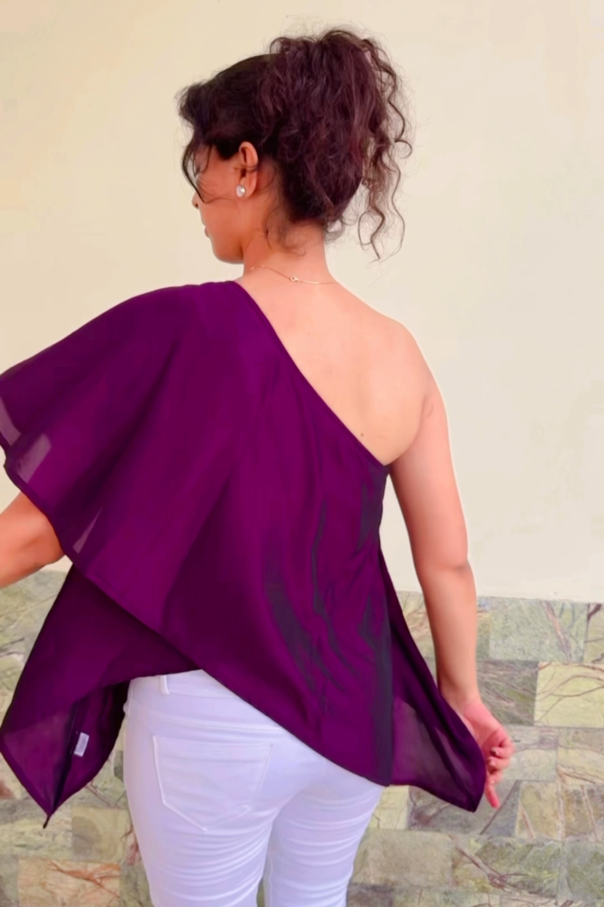 Person wearing a purple one-shoulder top and white pants against a neutral background
