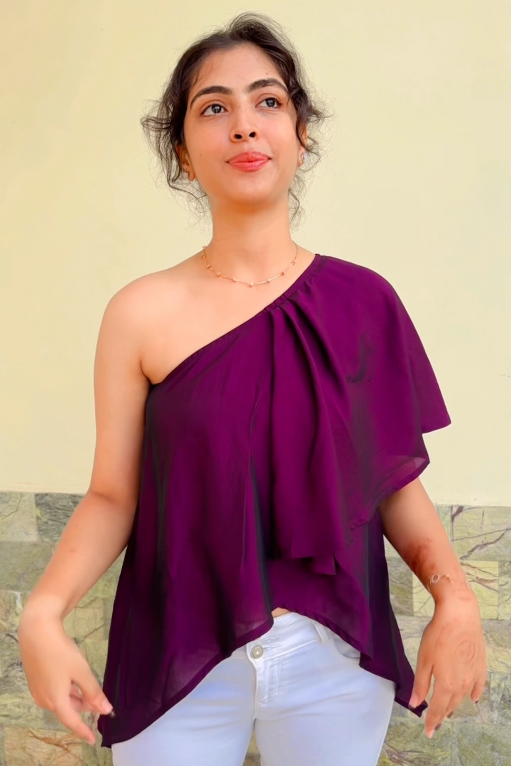 Woman wearing a one-shoulder purple top and white pants against a plain background