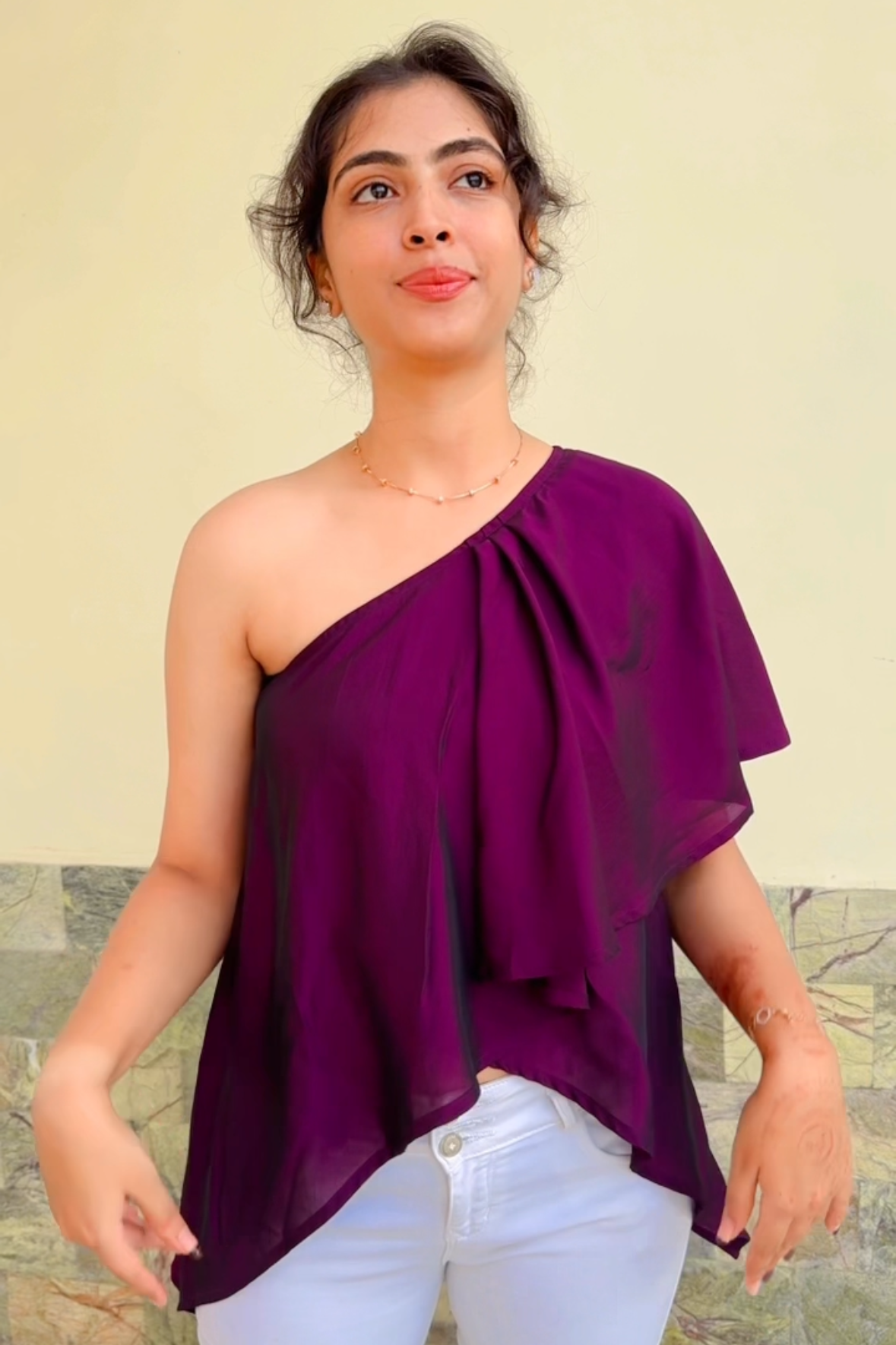 Woman wearing a one-shoulder purple top and white pants against a plain background