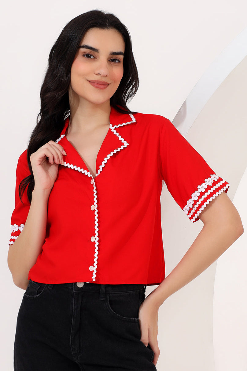 Woman in a red blouse with white trim standing in an outdoor setting with modern buildings and palm trees.