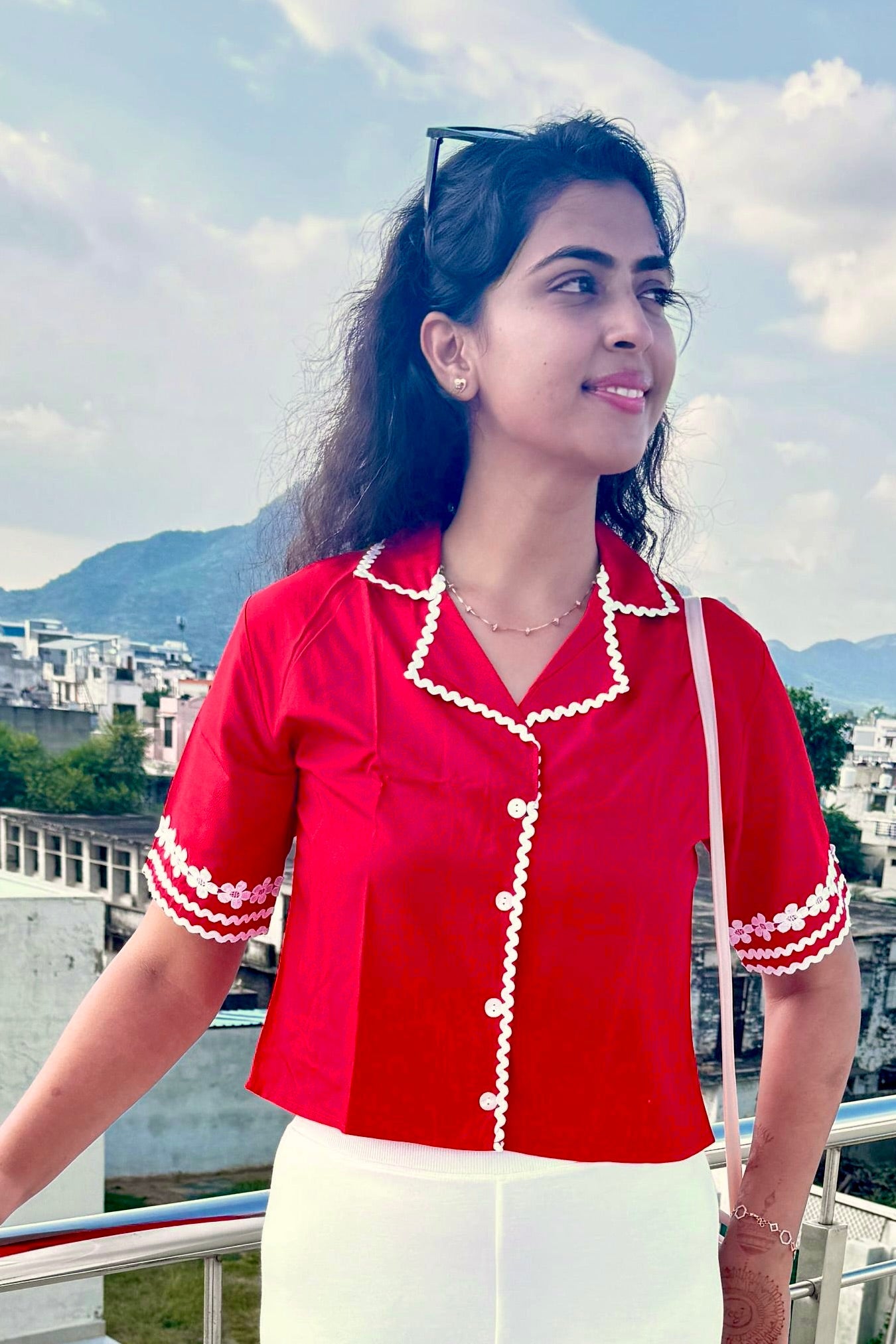 Woman wearing a red blouse with white trim on a balcony with a cityscape and mountains in the background