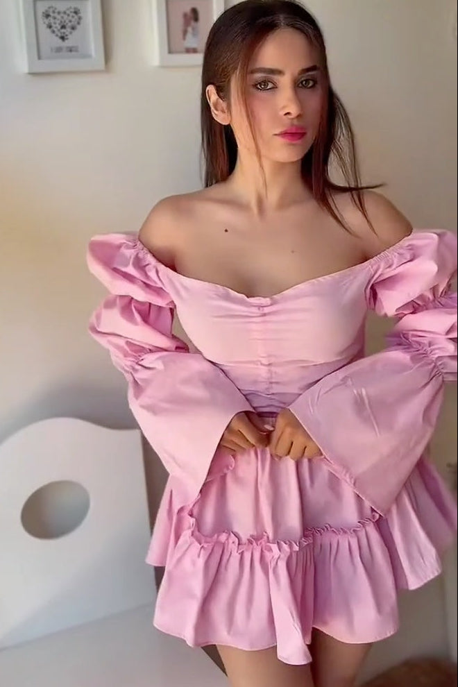 A woman wearing a light pink off-the-shoulder mini dress with dramatic puffed sleeves and a ruffled hem, standing indoors against a light-colored wall.