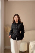 A woman wearing a black satin-like peplum blouse with a tie waist and white trousers, standing in a modern bedroom setting with beige walls, a bed, and a stool.