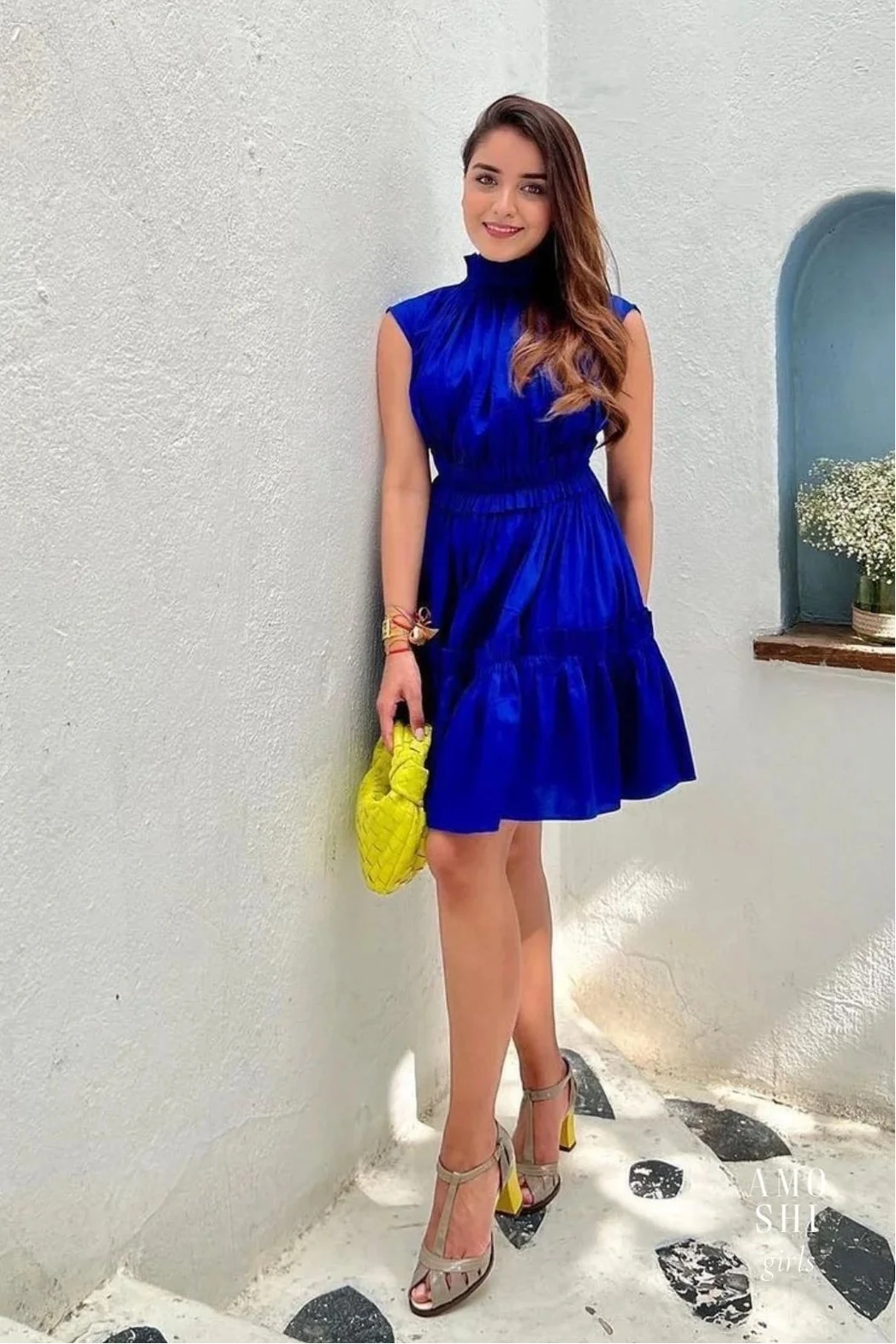 Woman in a blue dress holding a yellow bag against a white wall.