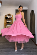 A woman wearing a bright pink sleeveless midi dress with a fitted bodice and a full layered tulle skirt, posing indoors with her hand lifting the skirt to show its volume. She is wearing sparkly silver high heels in a room with gray wood floors and neutral walls.