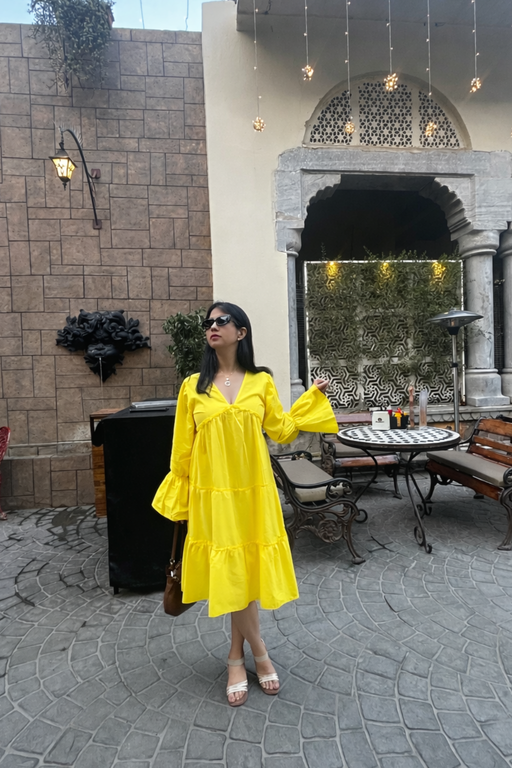 Woman in a yellow dress standing in an outdoor setting with a stone wall and patio furniture.