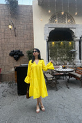Woman in a yellow dress standing in an outdoor setting with a stone wall and patio furniture.